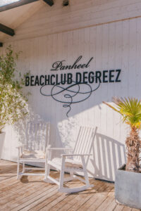 Ingang Beachclub Degreez met witte stoelen en logo op houten wand.