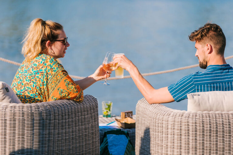 Man en vrouw proosten met een drankje op het terras aan het water bij Beachclub Degreez in Limburg.