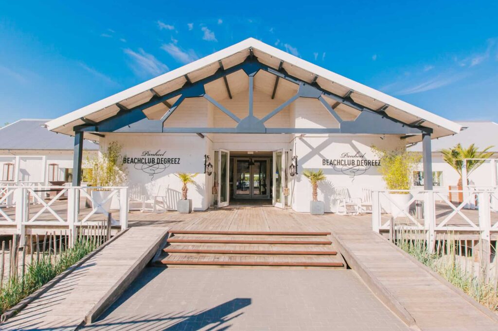 Vooraanzicht van Beachclub Degreez in Limburg met blauwe lucht, houten terras en tropische sfeer aan het water.