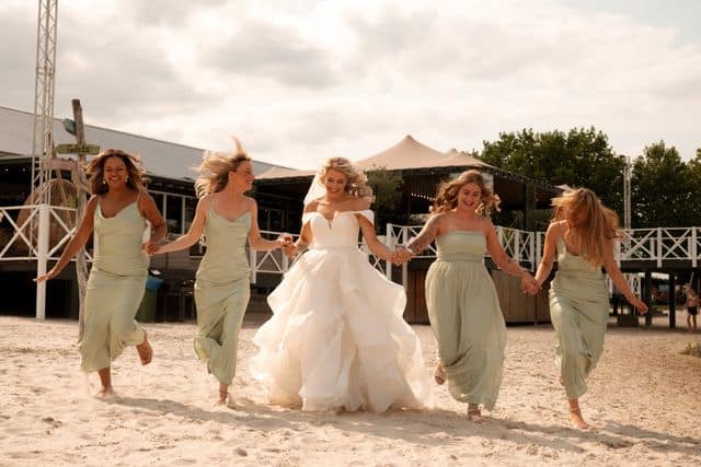 Bruid loopt samen met haar bruidsmeisjes over het strand bij Beachclub Degreez in Limburg.