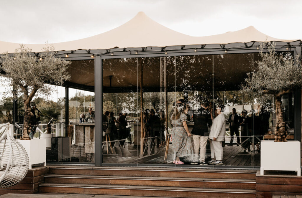 Het Waterfront van Beachclub Degreez met gasten achter glazen wanden en een terras aan het water.